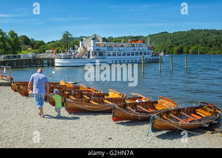 Ein Mann und ein kleiner Junge zu Fuß neben Ruderbooten auf das Ufer des Lake Windermere mit einem Tour-Dampfer im Hintergrund Stockfoto