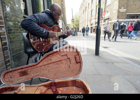 Eine Straße Straßenmusiker spielt Geld mit einer e-Gitarre für Spenden auf der Brick Lane in London während der 1. Mai Feiertag Stockfoto