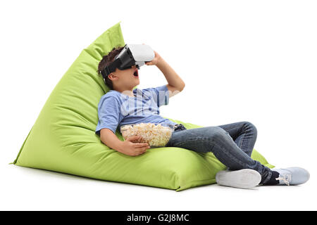 Kleiner Junge sucht in VR-Brille und Essen Popcorn sitzt auf einem Sitzsack isoliert auf weißem Hintergrund erstaunt Stockfoto