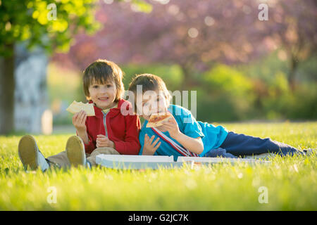 Zwei kleine Jungs mit Abendessen mit Pizza auf Sonnenuntergang, Picknick auf den Rasen-Frühling im blühenden Garten Stockfoto