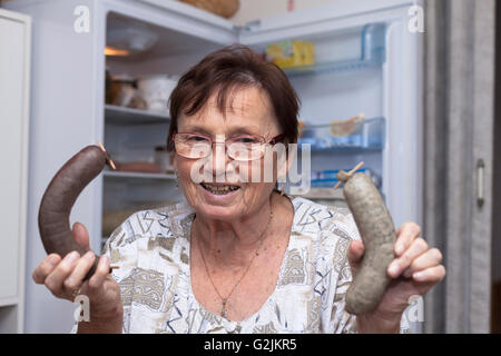 Beautiful senior Woman holding Schwein Leberwürste stehend vor der offenen Kühlschrank in der Küche. Stockfoto