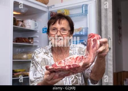 Glücklich senior Frau hält rohe Schweinerippchen vor den offenen Kühlschrank steht in der Küche. Stockfoto