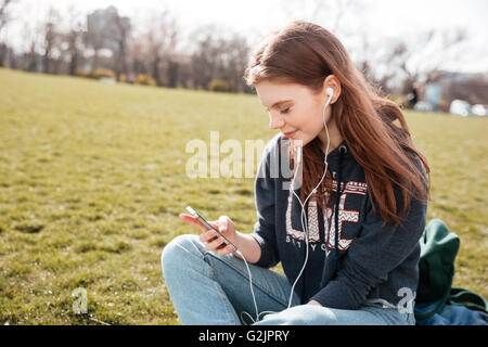 Niedlichen lächelnde junge Frau sitzt auf Rasen und nachdenklich aus Handy anhören Stockfoto