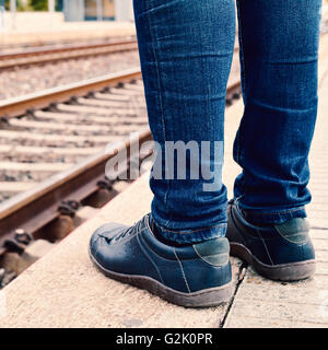 Nahaufnahme von den Füßen eines jungen Mannes trägt Jeans, der den Zug am Bahnsteig des Bahnhofs erwartet Stockfoto