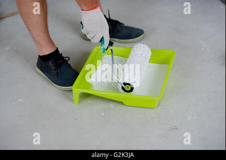 Auf einem grauen Betonboden ist eine gelbe Schale. In ihm weiße Farbe. Die Hand in einen Handschuh arbeiten taucht die Walze in Farbe. Stockfoto