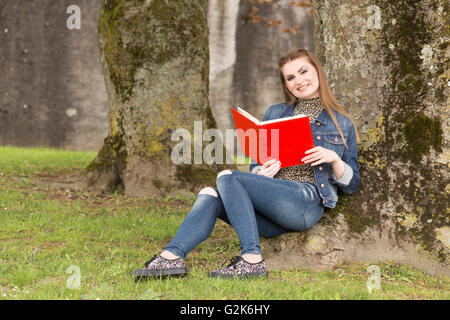 Eine junge braune dunkelhaarige Frau in blauen Jeans sitzt in einem Park und einem großen roten Buch. Stockfoto