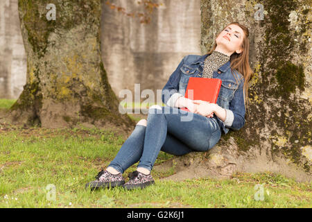 Eine junge braune dunkelhaarige Frau in blauen Jeans sitzt in einem Park und einem großen roten Buch. Stockfoto