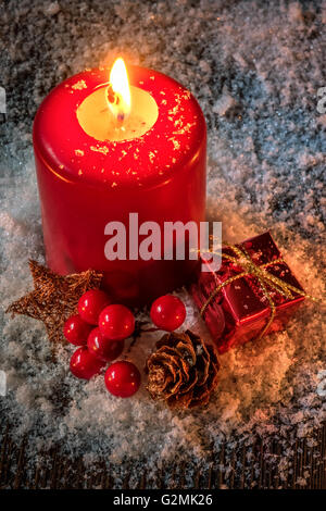 Atmosphärische Weihnachtskarte oder Hintergrund mit roten brennenden Kerze, Gegenwart, Tannenzapfen und Preiselbeeren auf einem Holztisch gedeckt w Stockfoto