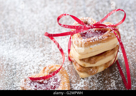 Frische Marmelade gefüllt Herzkekse mit Puderzucker und Band auf einem rustikalen Wodden Tisch. ISE es für ein Rezept oder eine Grußkarte Stockfoto