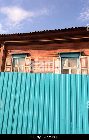 Alte Orange lackiert Holzhaus mit kunstvoll geschnitzten Fenstern versteckt durch einen großen grünen Metallzaun mit Sonne und blauer Himmel Stockfoto