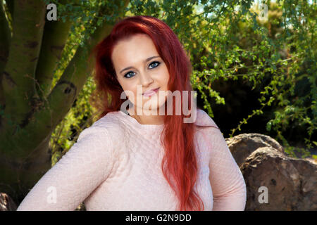 Eine schöne, Teenie-Alter Mädchen mit gefärbten roten Haaren posiert für ein Porträt vor Bäumen.  Sie hat einen geschlossenen Mund Lächeln. Stockfoto
