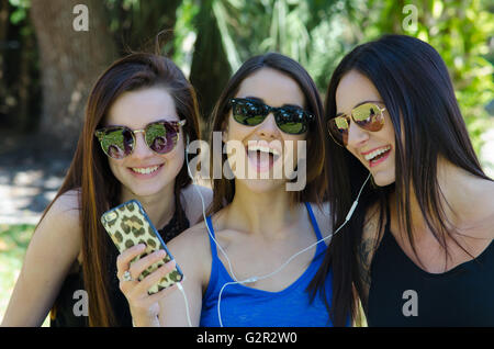 Drei junge Mädchen hören Sie Musik auf ihrem Handy lächelnd in einem Park in Sonnenbrillen Stockfoto