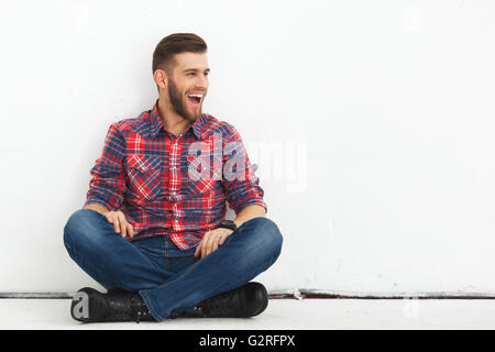 Porträt von glücklich überrascht hübschen jungen Mann in Freizeitkleidung gegen weiße Wand sitzen. Stockfoto