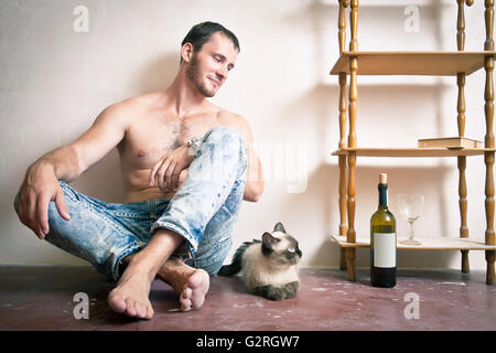 Ein Mann mit einer Flasche Wein in der Hand auf dem Boden in der Nähe der Katze sitzend Stockfoto