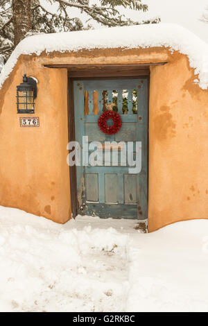 Eine alte Holztür mit einem roten Chili-Kranz auf einem Adobe-Stil Haus im Stadtteil Arroyo Tierra Blanca nach einem Winter Schneefall 13. Dezember 2015 in Santa Fe, New Mexico. Stockfoto