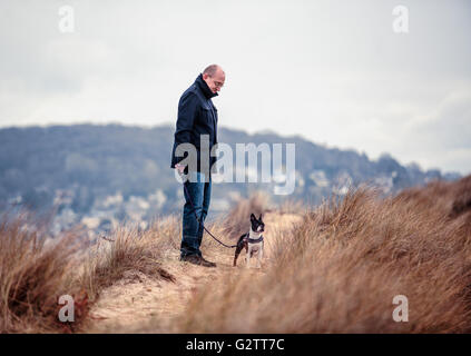 Mann spielt mit agilen schwarz-weiß Boston Terrier an einem Sandstrand in der Normandie an einem sonnigen Tag Stockfoto