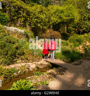 Zwei kleine Mädchen im roten Mantel auf einer Brücke über einen Bach in einem Garten Stockfoto