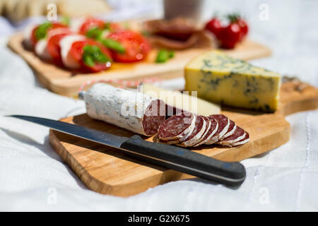 Nahaufnahme der geschnittene Salami in ein Picknick mit Tomatensalat mit Mozzarella, Parmaschinken, Erdbeeren, Oliven und Espressokanne in einem Park. Stockfoto