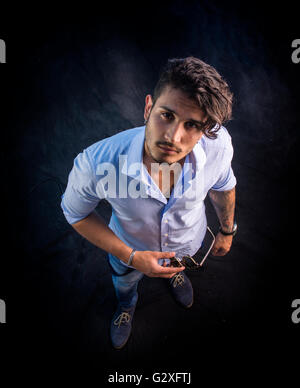 Porträt von Brünette junge Mann in hellblaues Hemd und Jeans, stehend im Studio gedreht von oben Perspektive gegen dunkle backg Stockfoto