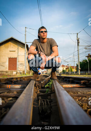 Attraktive junge Mann sitzen auf Eisenbahn, graues T-shirt und Jeans tragen, wegschauen Stockfoto