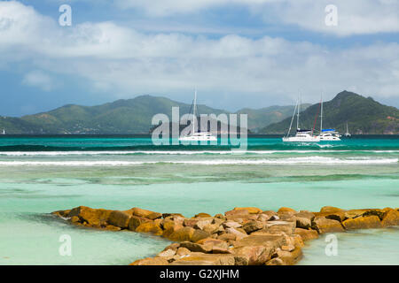 Weißer Traumstrand in La Digue, Seychellen, geschützt durch eine Reihe von Felsbrocken führt ins Bild, einige Boote im Hintergrund Stockfoto