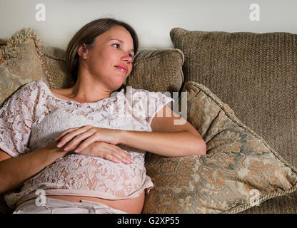 nachdenkliche schwangere Frau auf einem Sofa sitzt und streichelte ihren Bauch Stockfoto