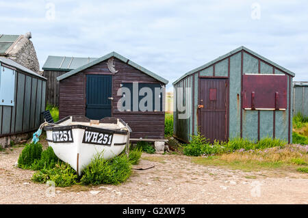 Eine weiße Fischerboot ist vor einer braunen Holz Strandhütte mit grünen Pflanzen wachsen aus dem steinigen Boden gebunden Stockfoto