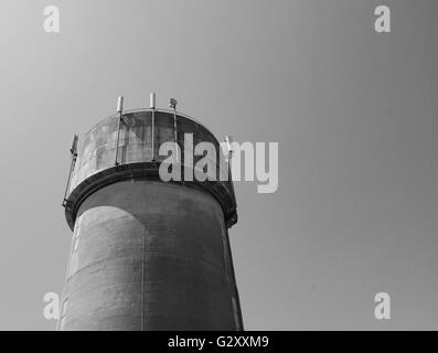 Stark verwitterter Wasserturm aus Gussbeton, der vor einem klaren blauen Himmel gesehen wird. Auf dem Turm sind neu installierte 5G Telecom Masten zu sehen. Stockfoto