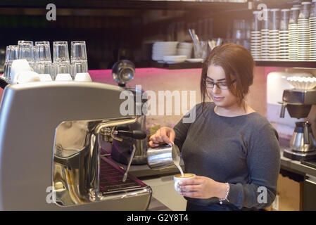 Weibliche Barista oder Kellnerin mache eine Tasse Cappuccino in einem Kaffeehaus oder in der Cafeteria der heiße aufgeschäumte Milch in den Kaffee gießen Stockfoto