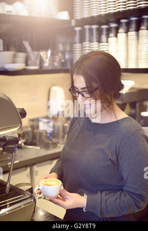 Weibliche Barista oder Kellnerin hält eine Tasse Cappuccino in einem Kaffeehaus oder in der Cafeteria, lächelnd zeigt Zufriedenheit Stockfoto