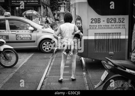 Mutter halten zwei Kleinkinder überqueren einer viel befahrenen Straße in Hanoi, Vietnam. Stockfoto