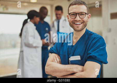 Optimistische junge männliche Krankenhaus Chirurg stehend vor seinen Kollegen mit verschränkten Armen strahlt in die Kamera Lächeln Stockfoto