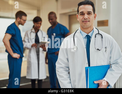 Zuversichtlich junger Hispanic-Arzt führt eine multirassische Ärzteteam steht im Vordergrund mit einer Datei unter seinem Arm lächelnd Stockfoto