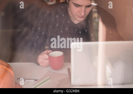 Warten auf Inspiration. Nachdenklicher junger Mann sitzt an seinem Arbeitsplatz, trinken Kaffee, Reflexion im Glas Stockfoto