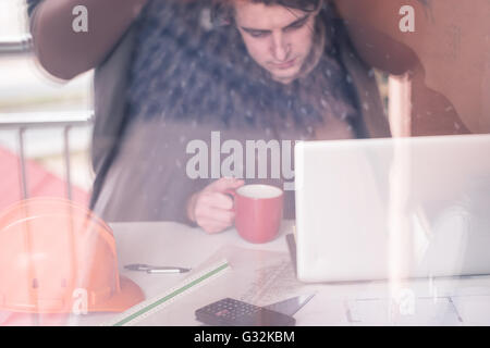 Warten auf Inspiration. Nachdenklicher junger Mann sitzt an seinem Arbeitsplatz, trinken Kaffee, Reflexion im Glas Stockfoto