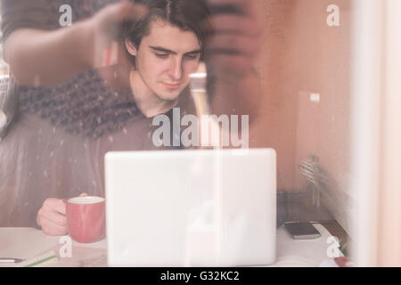 Warten auf Inspiration. Nachdenklicher junger Mann sitzt an seinem Arbeitsplatz, trinken Kaffee, Reflexion im Glas Stockfoto