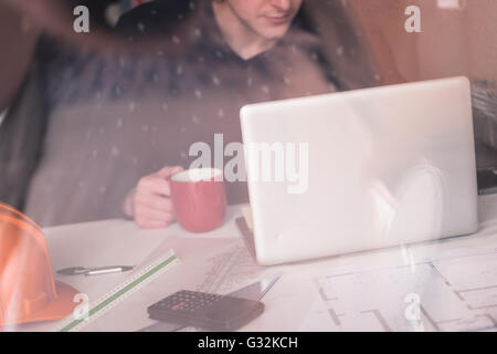 Warten auf Inspiration. Nachdenklicher junger Mann sitzt an seinem Arbeitsplatz, trinken Kaffee, Reflexion im Glas Stockfoto