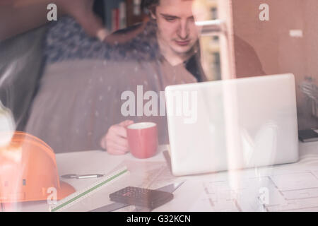Warten auf Inspiration. Nachdenklicher junger Mann sitzt an seinem Arbeitsplatz, trinken Kaffee, Reflexion im Glas Stockfoto