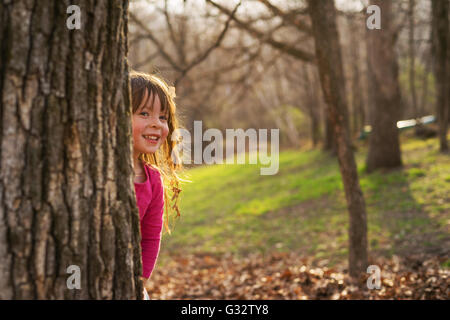 Lächelndes Mädchen versteckt sich hinter einem Baum im park Stockfoto
