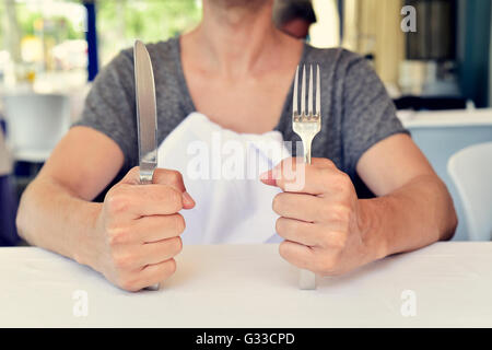 Closeup von einem kaukasischen Jüngling sitzt an einem Tisch warten auf das Essen, mit einem Messer in der Hand und einer Gabel in der anderen hand Stockfoto