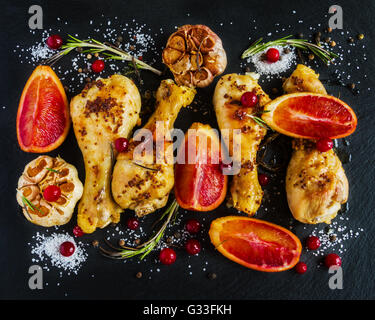 Gebratene Hähnchenschenkel, rote Orangen, Preiselbeeren, Knoblauch und Rosmarin auf dem schwarzen Hintergrund. Ansicht von oben. Stockfoto