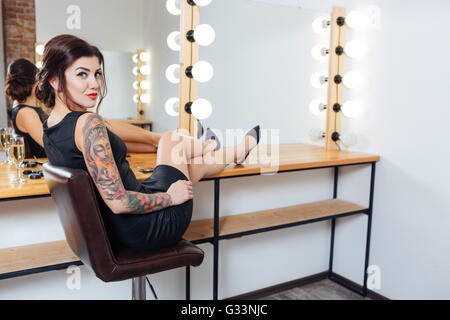 Glücklich tätowierte junge Frau im schwarzen Kleid und Schuhe im Ankleidezimmer mit Händen am Tisch sitzen Stockfoto