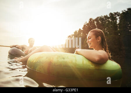 Junge Frau in einem Innertube bei Sonnenuntergang an einem See mit Mann im Hintergrund schweben. Paar genießt ihren Urlaub am See. Stockfoto