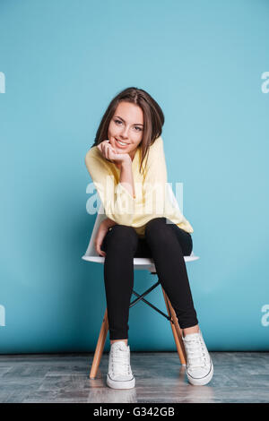 Junge Frau sitzt auf der Stuhl unter Kinn auf dem blauen Hintergrund isoliert Stockfoto