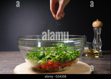 Hand, die Zugabe von Salz, Gemüsesalat. Schüssel mit frischen grünen Salat, Tomaten, Gurken auf Holztisch vor einem dunklen Hintergrund. Stockfoto