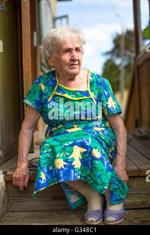 Alte Frau sitzt auf der Veranda des ländlichen Hauses. Stockfoto