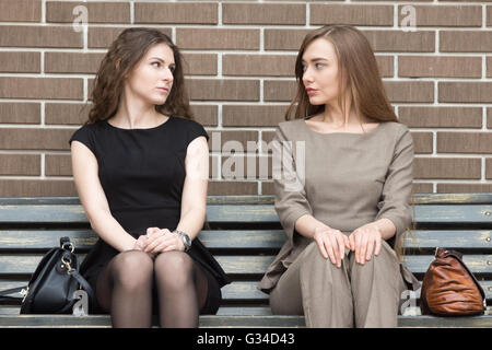 Porträt von zwei schönen jungen Frau Rivalen auf Bank sitzen nebeneinander und sahen einander mit anspruchsvollen Stockfoto