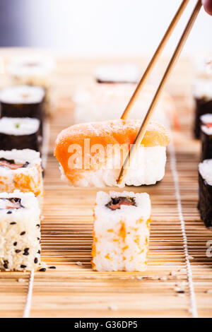 ein Lachs Nigiri Sushi abgeholt mit Stäbchen mit verschiedenen Arten von Maki-Sushi-Stücke auf einer hölzernen Sushi-Matte in der bac Stockfoto