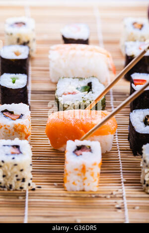 ein Lachs Nigiri Sushi abgeholt mit Stäbchen mit verschiedenen Arten von Maki-Sushi-Stücke auf einer hölzernen Sushi-Matte in der bac Stockfoto