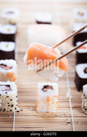 ein Lachs Nigiri Sushi abgeholt mit Stäbchen mit verschiedenen Arten von Maki-Sushi-Stücke auf einer hölzernen Sushi-Matte in der bac Stockfoto